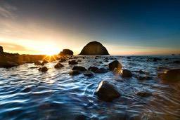 The sea is calm at sunrise, the rocks in the distance are bathed in golden light, and black rocks are scattered on the beach. - free wallpaper image