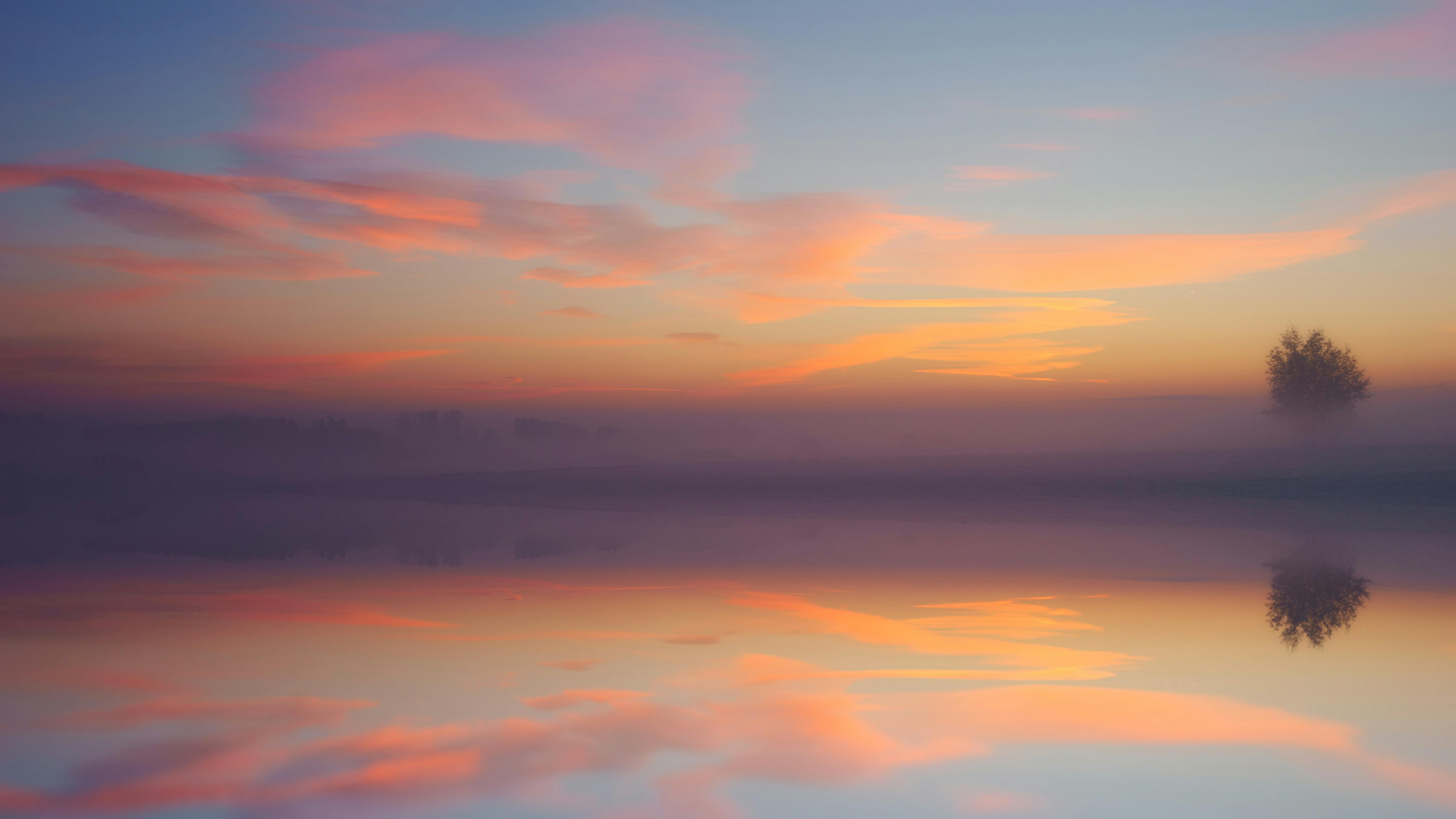 The sky is filled with pink and orange clouds at sunrise, the colors are reflected in the lake, and there is a tree in the distance. - free wallpaper image