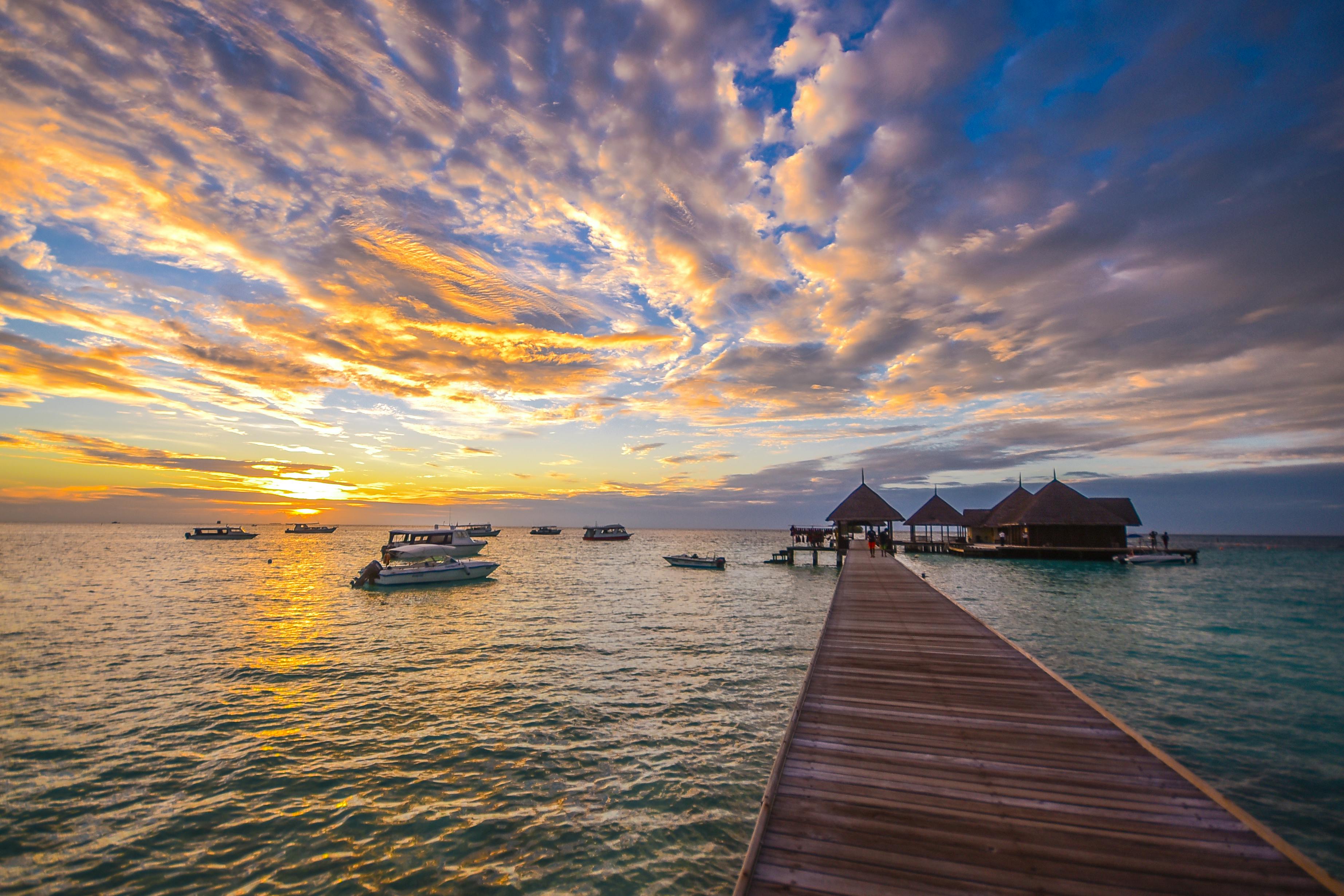 The sun sets over the sea, several yachts float on the water, a wooden pier leads to a resort hotel in the distance, surrounded by calm waters. - free wallpaper image