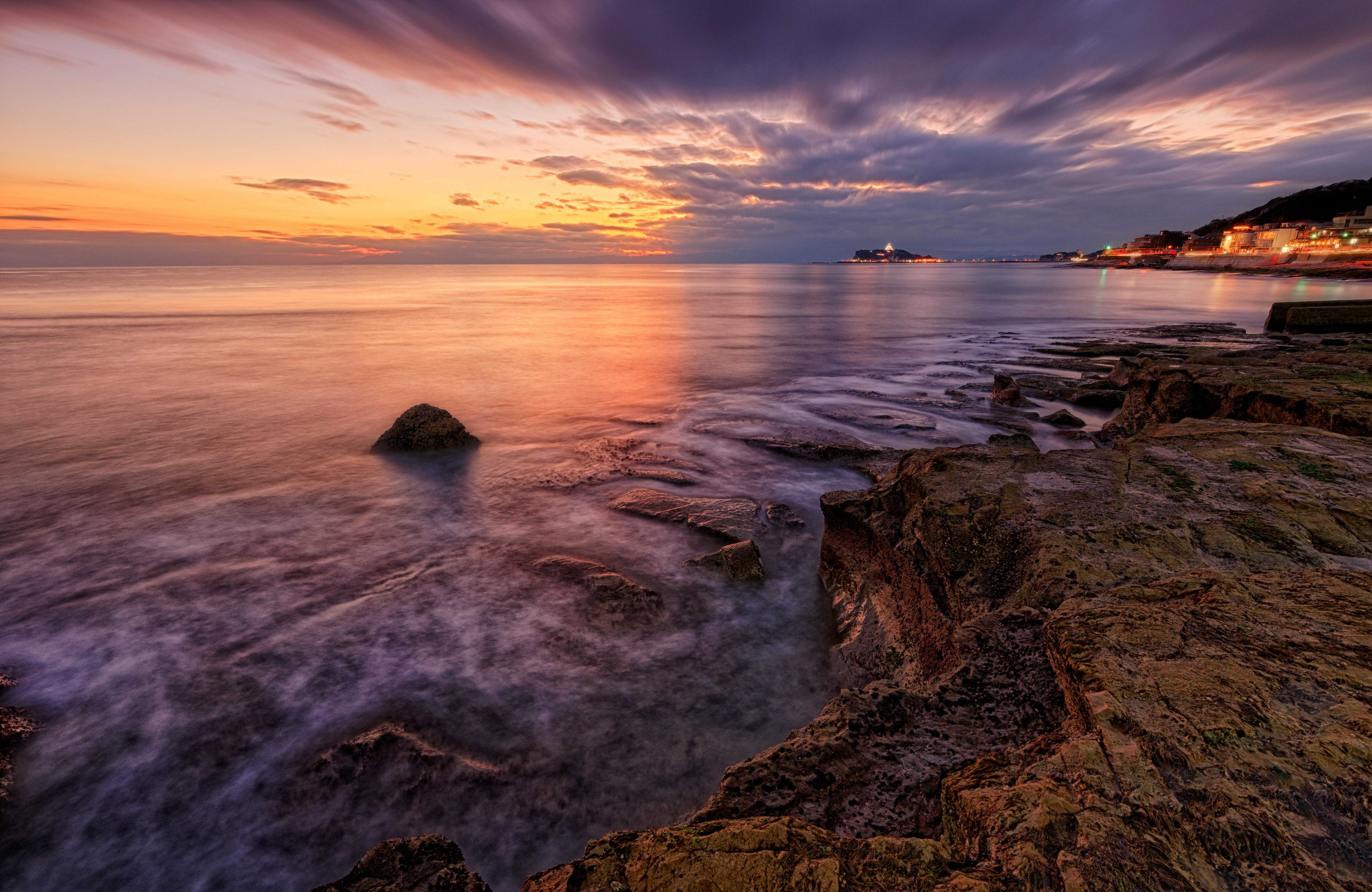 The sunset casts a glow over the rocky shoreline, with lights twinkling along the coast in the distance. The scene is breathtakingly beautiful. - free wallpaper image