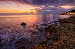 The sunset casts a glow over the rocky shoreline, with lights twinkling along the coast in the distance. The scene is breathtakingly beautiful. - free wallpaper image