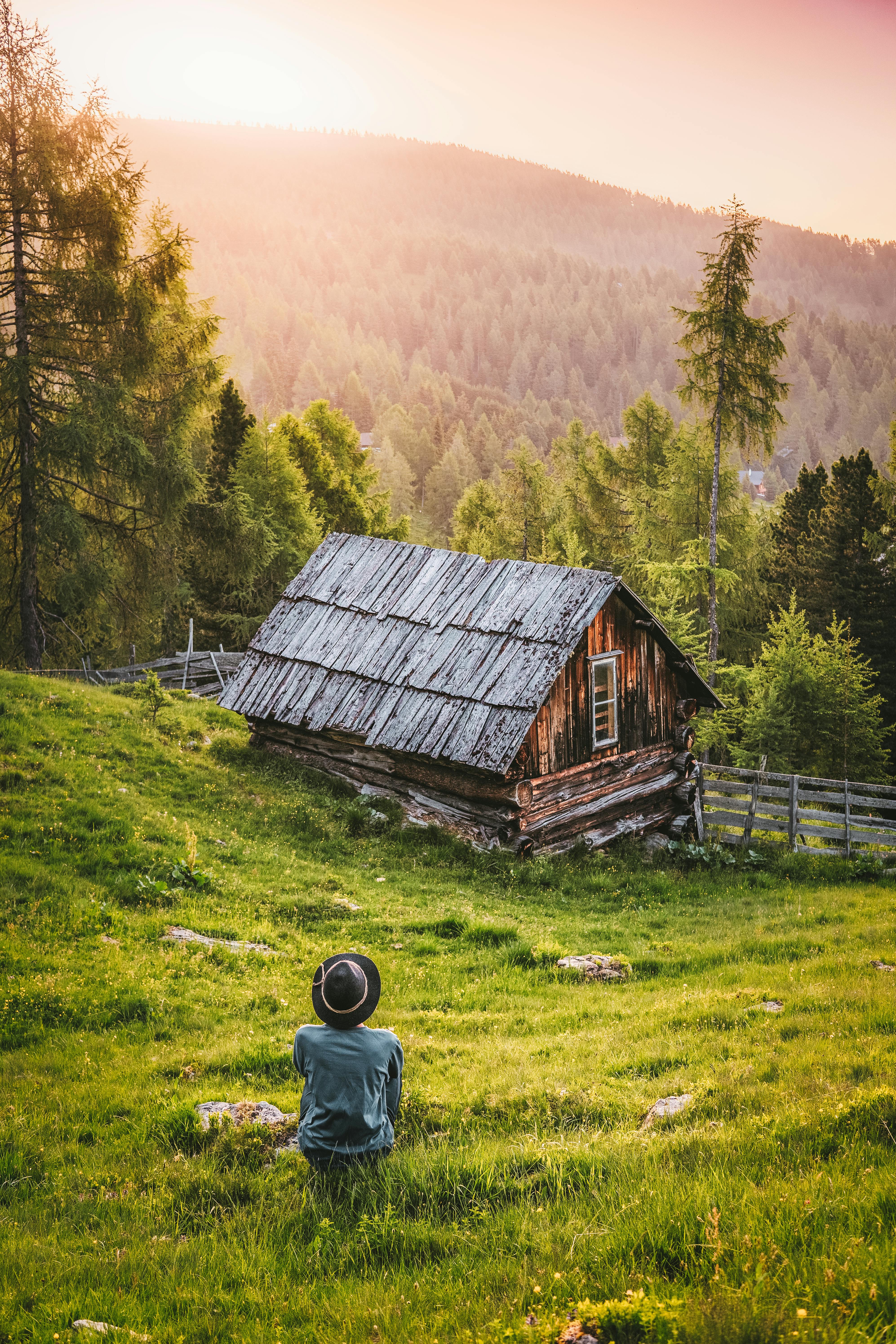 A person sits on a hillside under the setting sun, looking at a wooden cabin in the distance, enjoying the peace of nature. - free wallpaper image