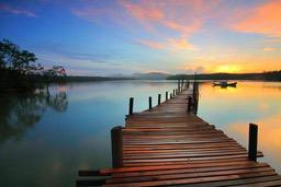 A wooden pier extends into a calm lake with a golden sunset in the distance and a cloudy sky. - free wallpaper image