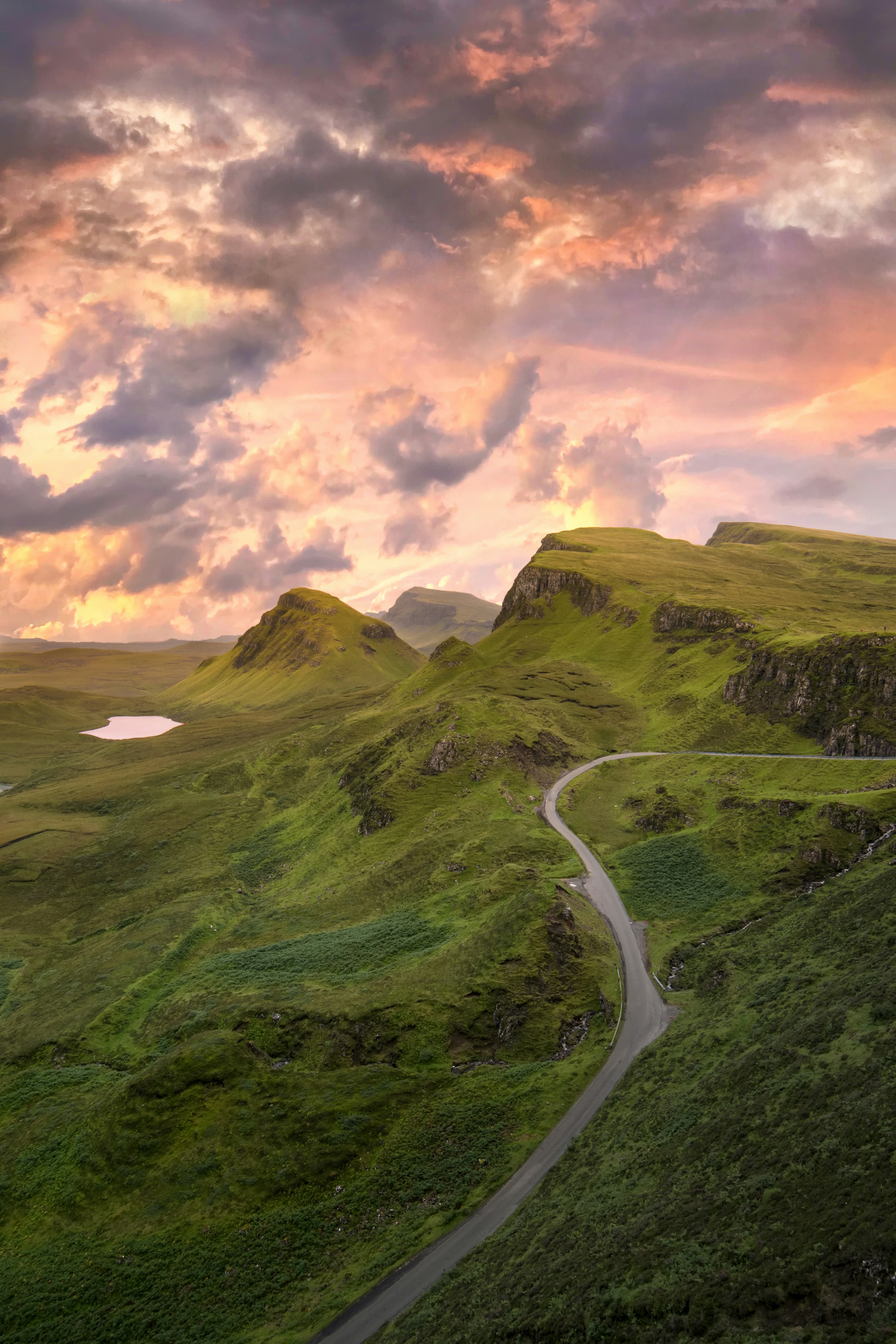 The setting sun casts a warm glow over rolling green hills as a winding road snakes its way through the landscape. - free wallpaper image