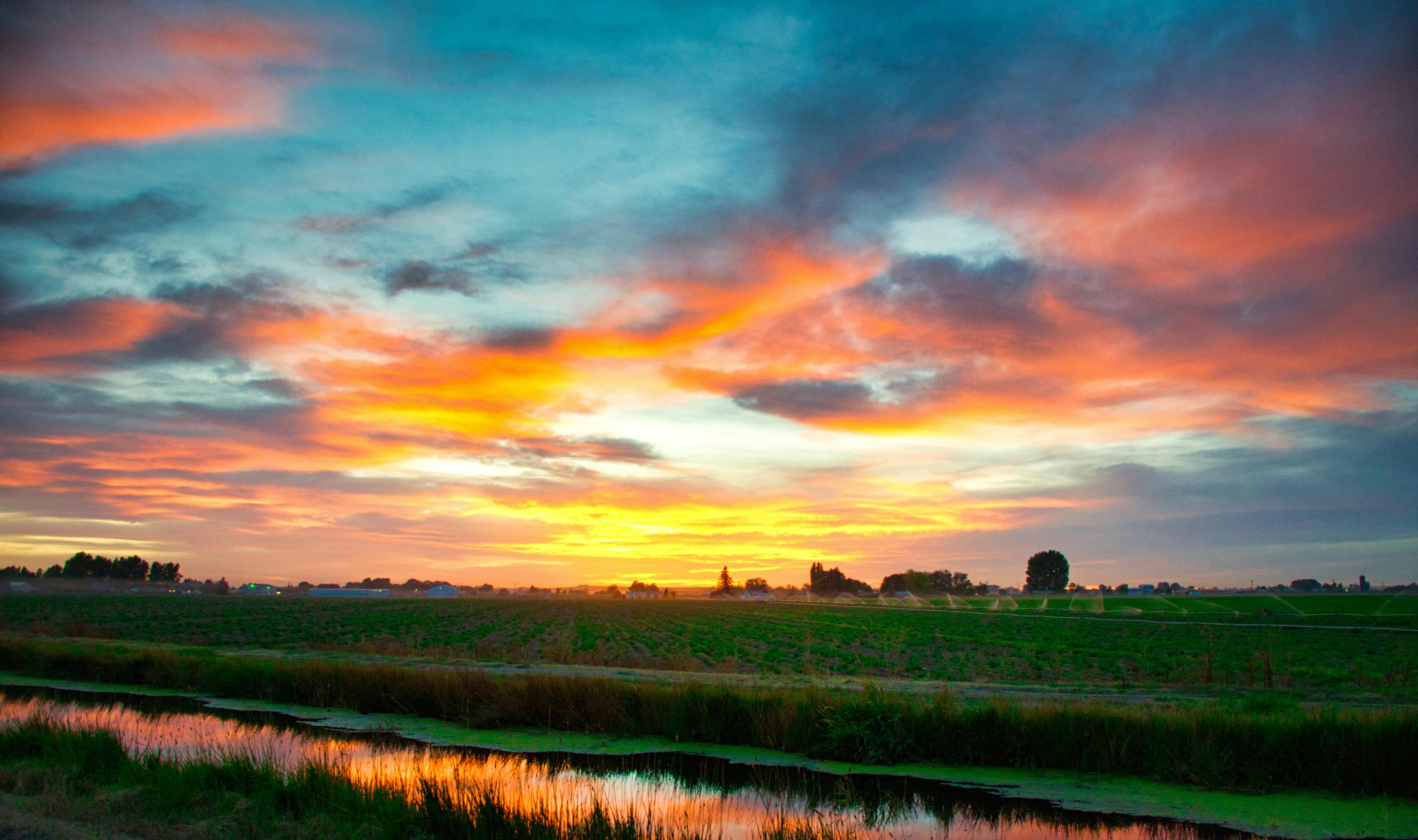 The sky is painted orange and blue as the sun sets, there is a river in the distance with green grass along the banks. - free wallpaper image