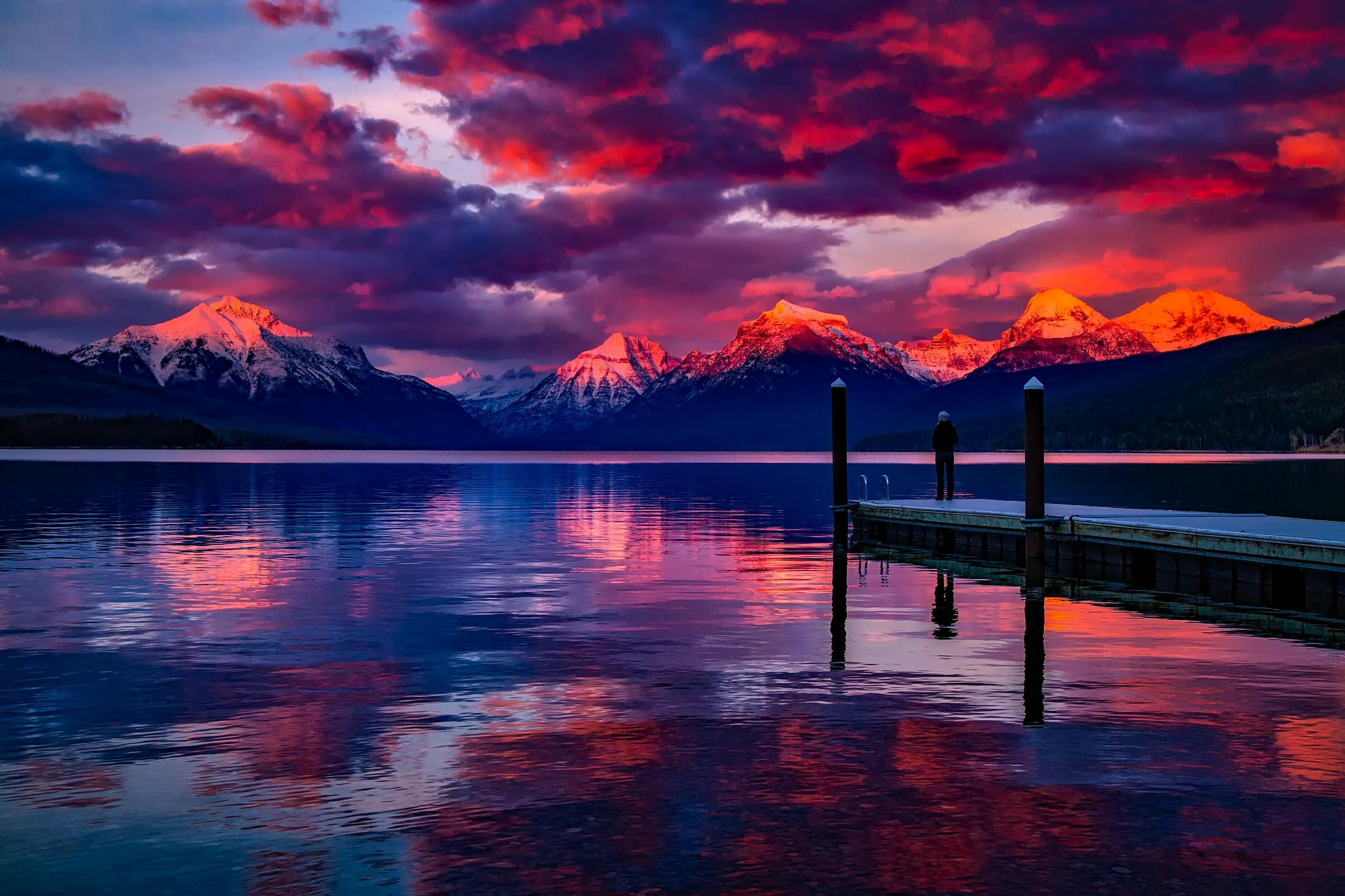 The sunset paints the sky with vibrant colors, reflecting off the calm lake, while a lone figure stands on a dock, taking in the beauty of the mountain range. - free wallpaper image