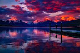 The sunset paints the sky with vibrant colors, reflecting off the calm lake, while a lone figure stands on a dock, taking in the beauty of the mountain range. - free wallpaper image