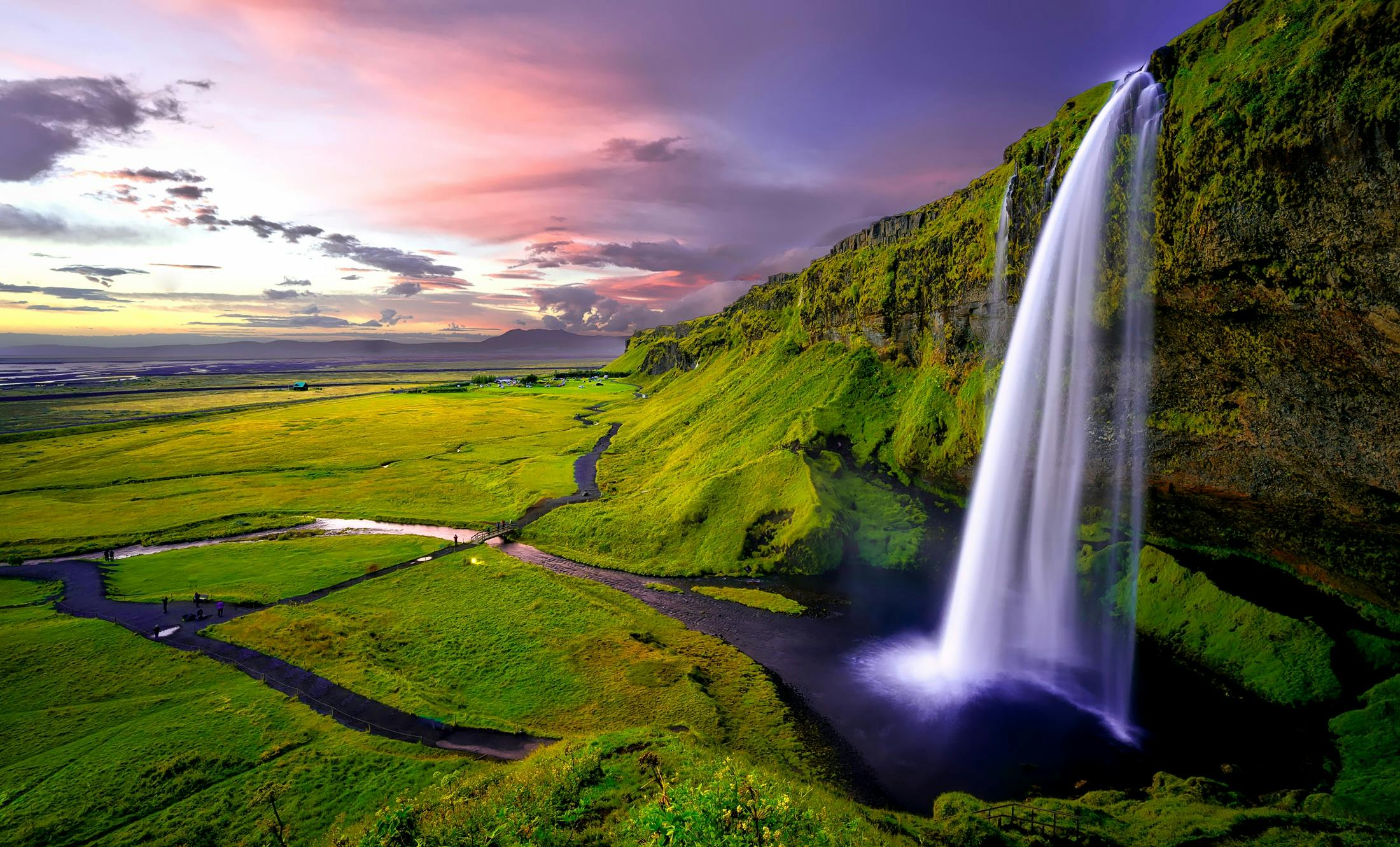 A waterfall cascades down a cliff in the setting sun, surrounded by green grass and a river, with distant mountains in the background. - free wallpaper image