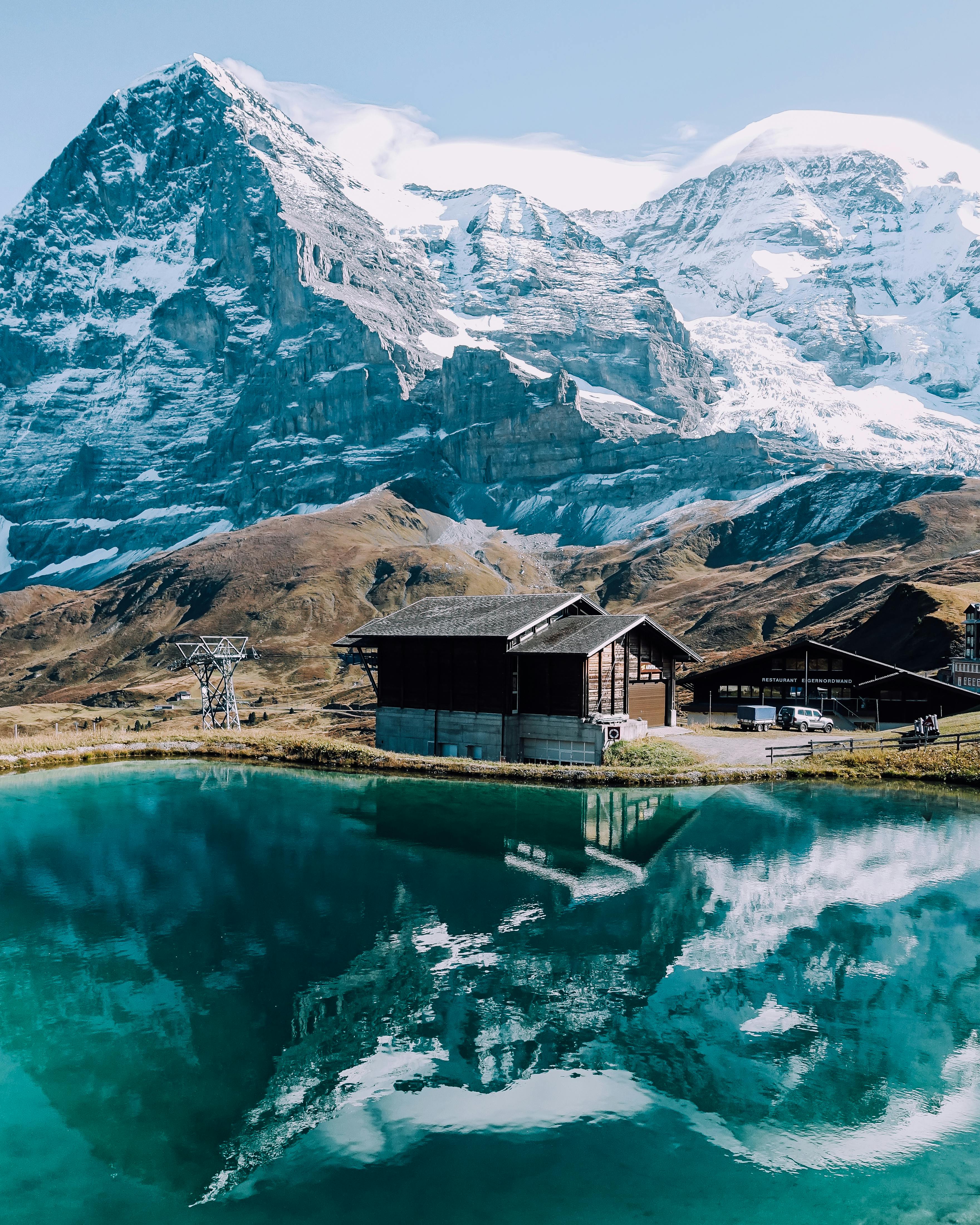 A wooden hut stands quietly by the lake under the snowy mountains, reflected in the clear lake water, forming a peaceful picture. - free wallpaper image