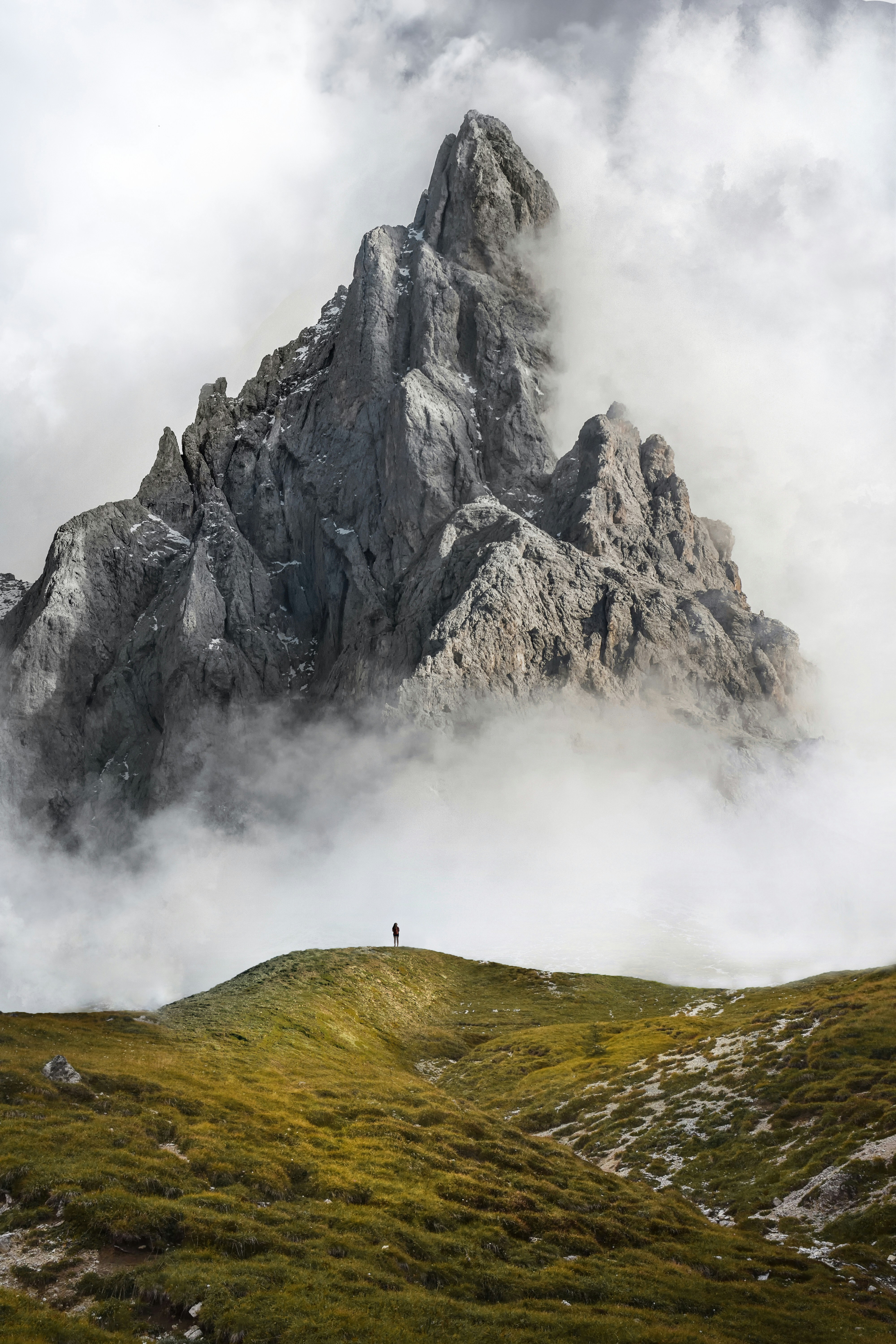 A towering mountain peak stands amidst the clouds, its summit obscured by fog, a green meadow at the foot of the mountain, a tiny figure standing on the hillside. - free wallpaper image