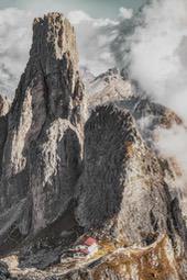 A towering rock peak stands amidst the cloudy mountains, with a small hut at the foot of the mountain. - free wallpaper image