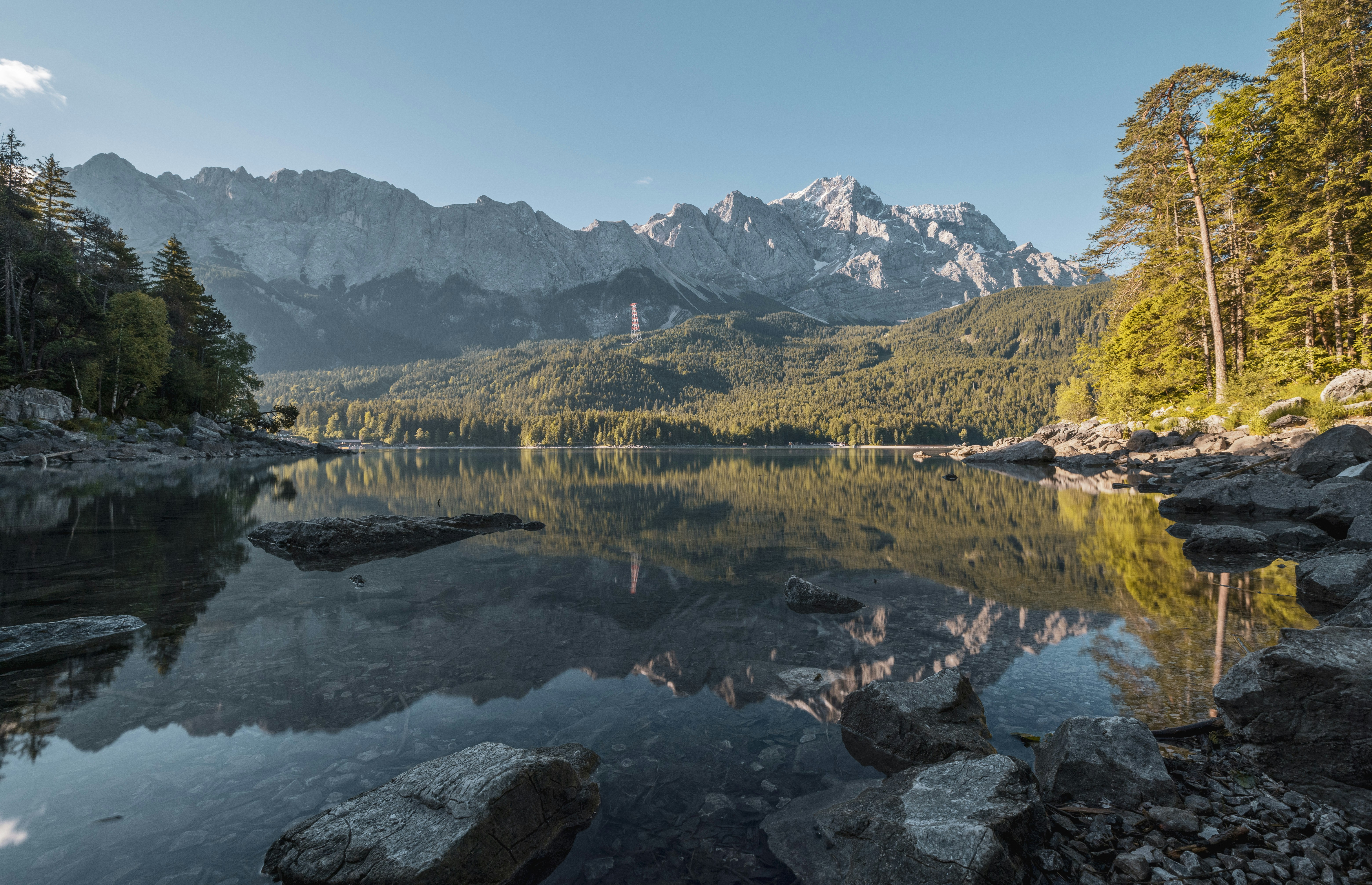 A tranquil lake reflects the towering mountains, with lush forest and scattered rocks along the shore. - free wallpaper image