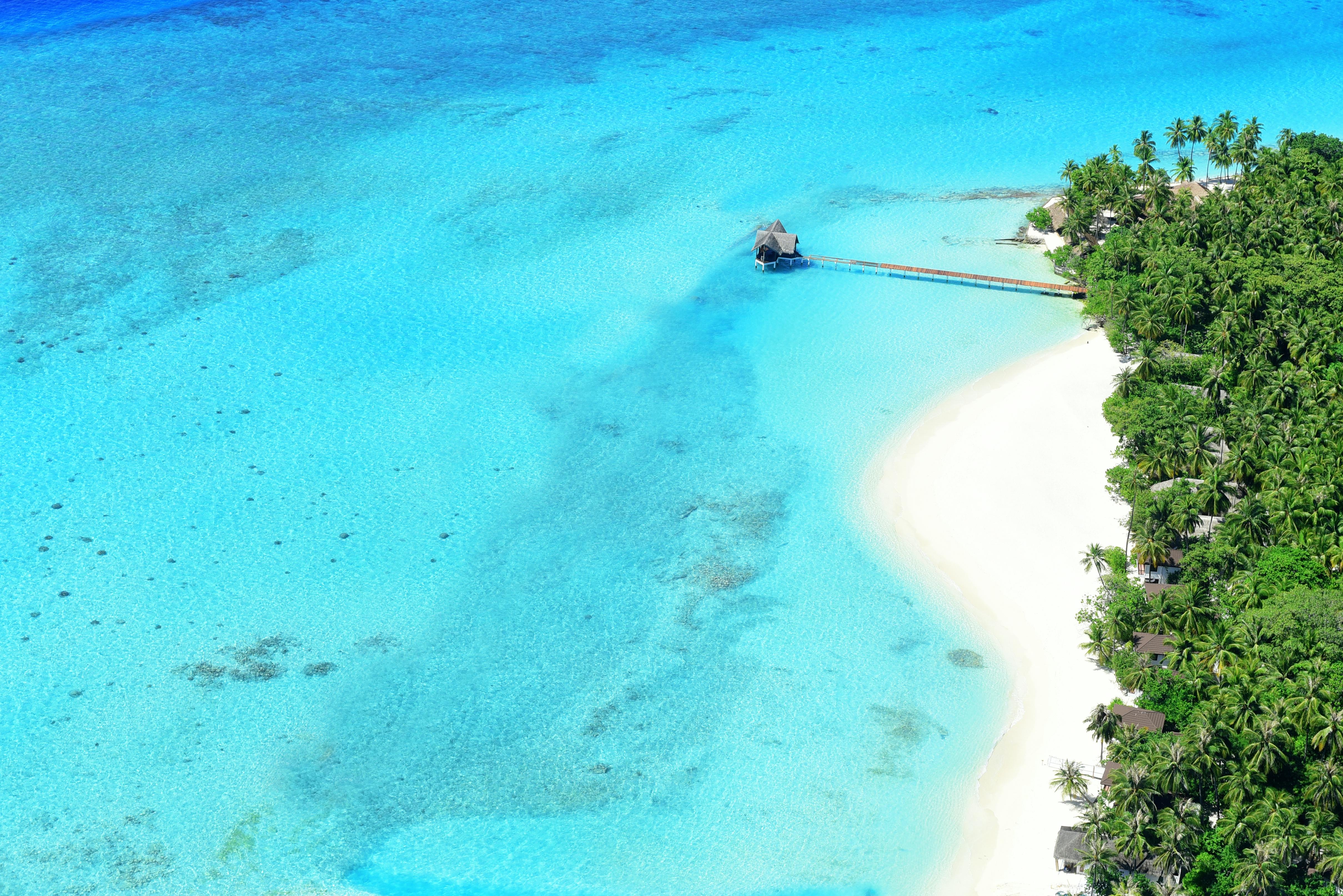 An aerial view of a pristine beach with clear blue water, white sand and lush green trees. There is a wooden structure on the beach, with a wooden bridge extending from it. - free wallpaper image