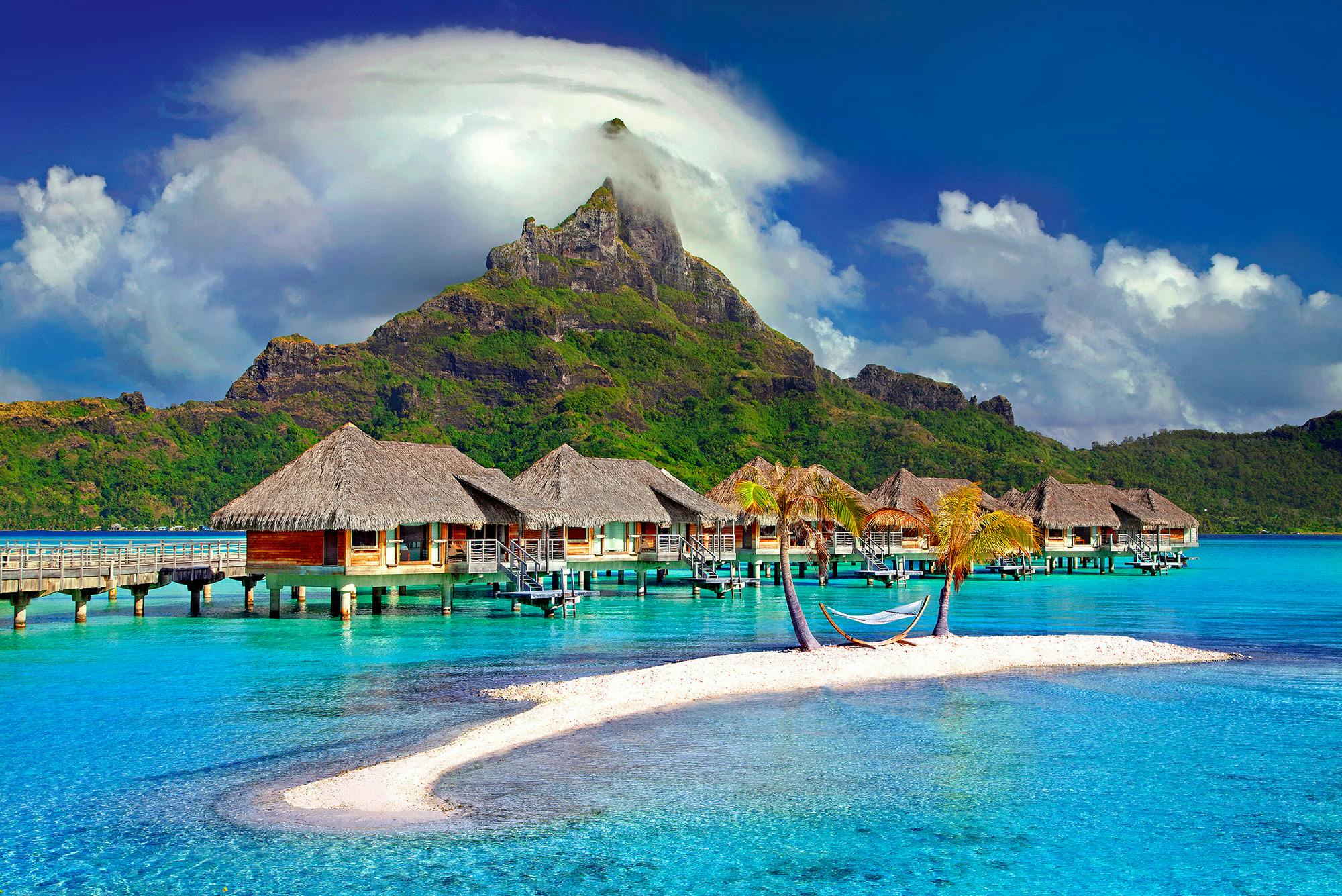 A row of wooden houses on the water are built in the azure water, next to a sandy beach with a hammock on it. There are lush mountains in the distance. - free wallpaper image