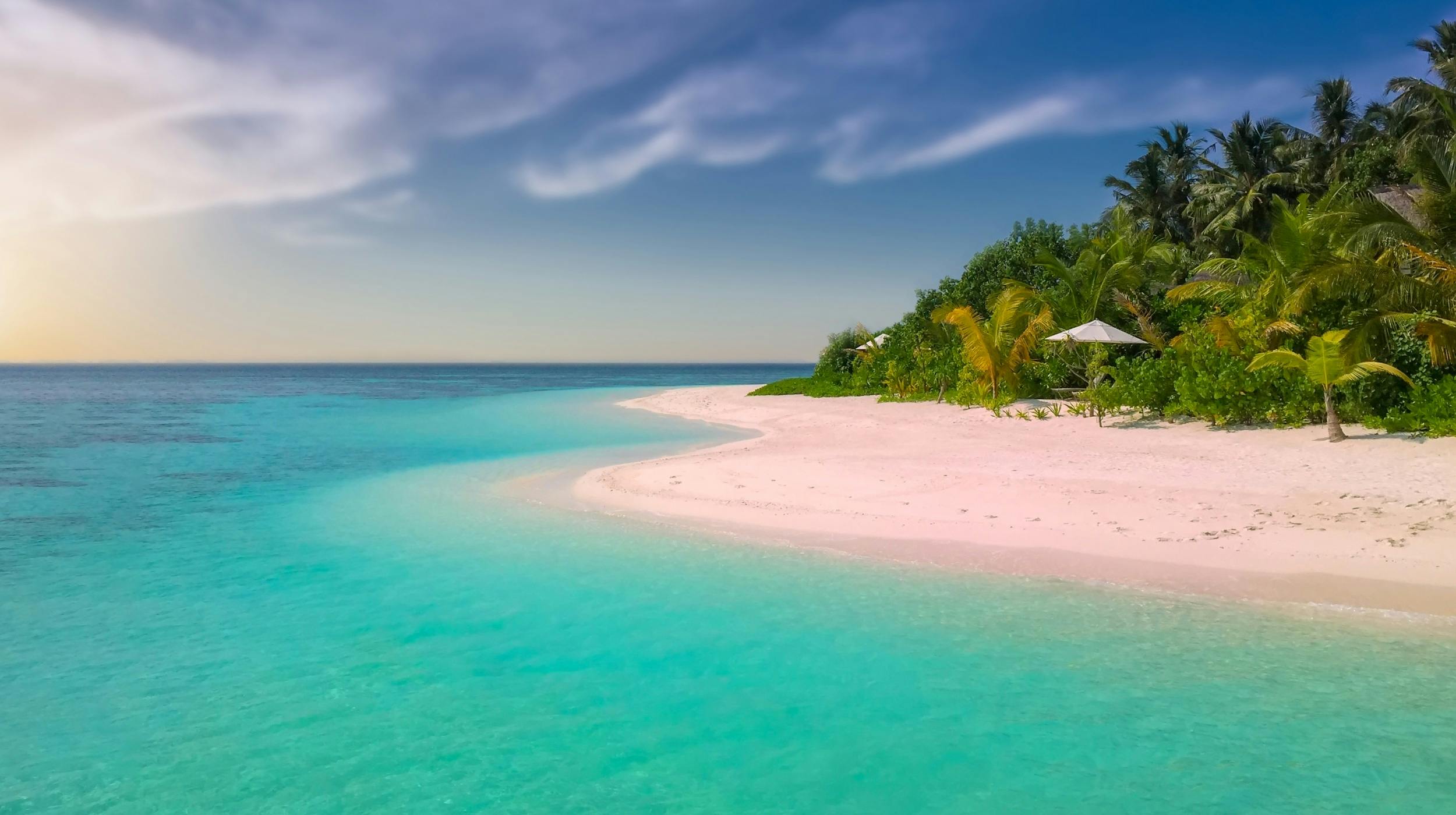 A tropical beach with white sand, clear water, lush greenery on the shore, and a blue sky in the distance. - free wallpaper image