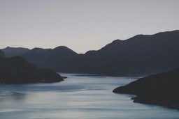 Mountains are layered against a twilight sky, a still body of water reflects the sky's hues and there are a few houses in the distance. - free wallpaper image