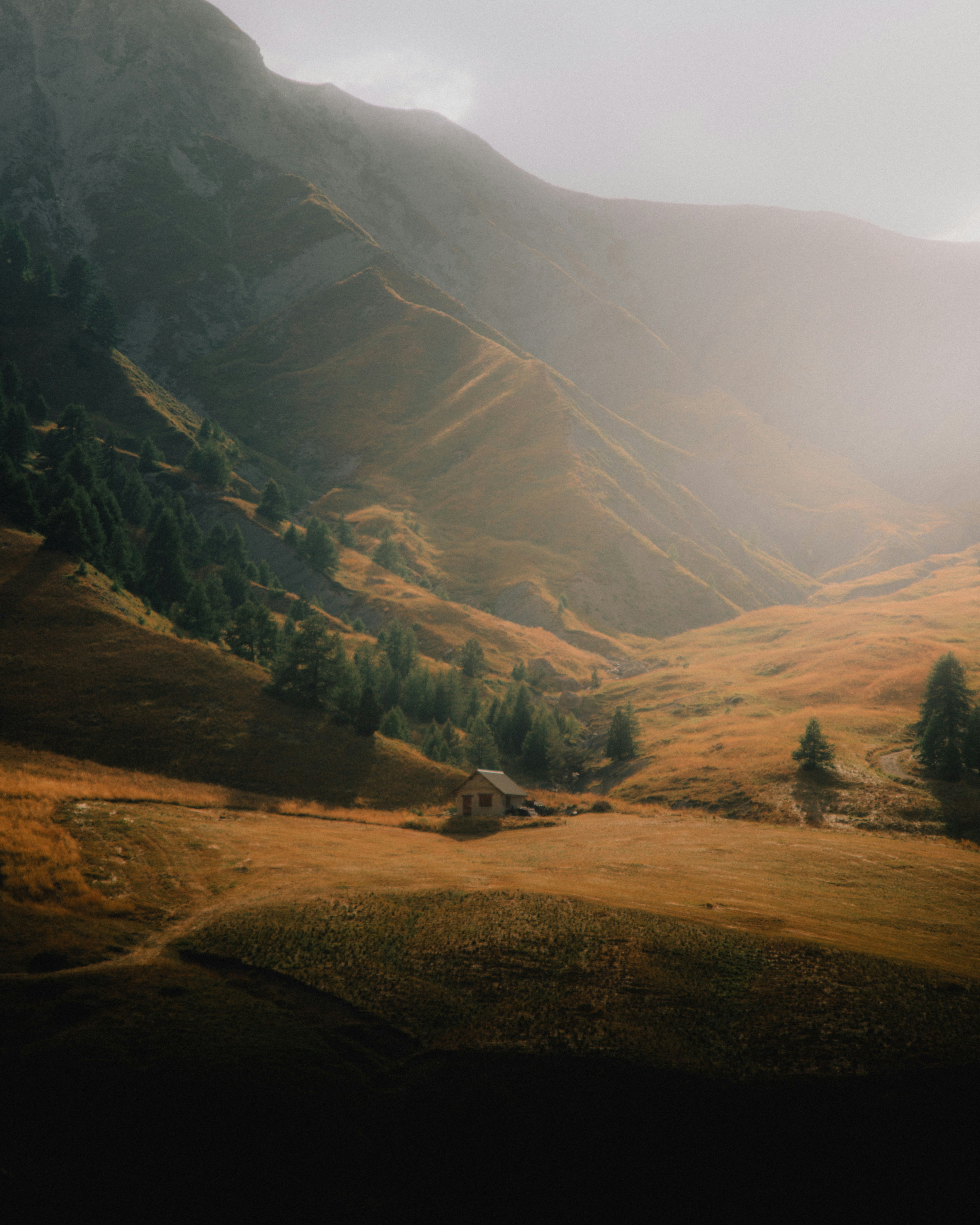 A small cabin sits on a green hillside in a valley, with grey mountains in the distance and sunlight illuminating the sky. The scene is peaceful and serene. - free wallpaper image