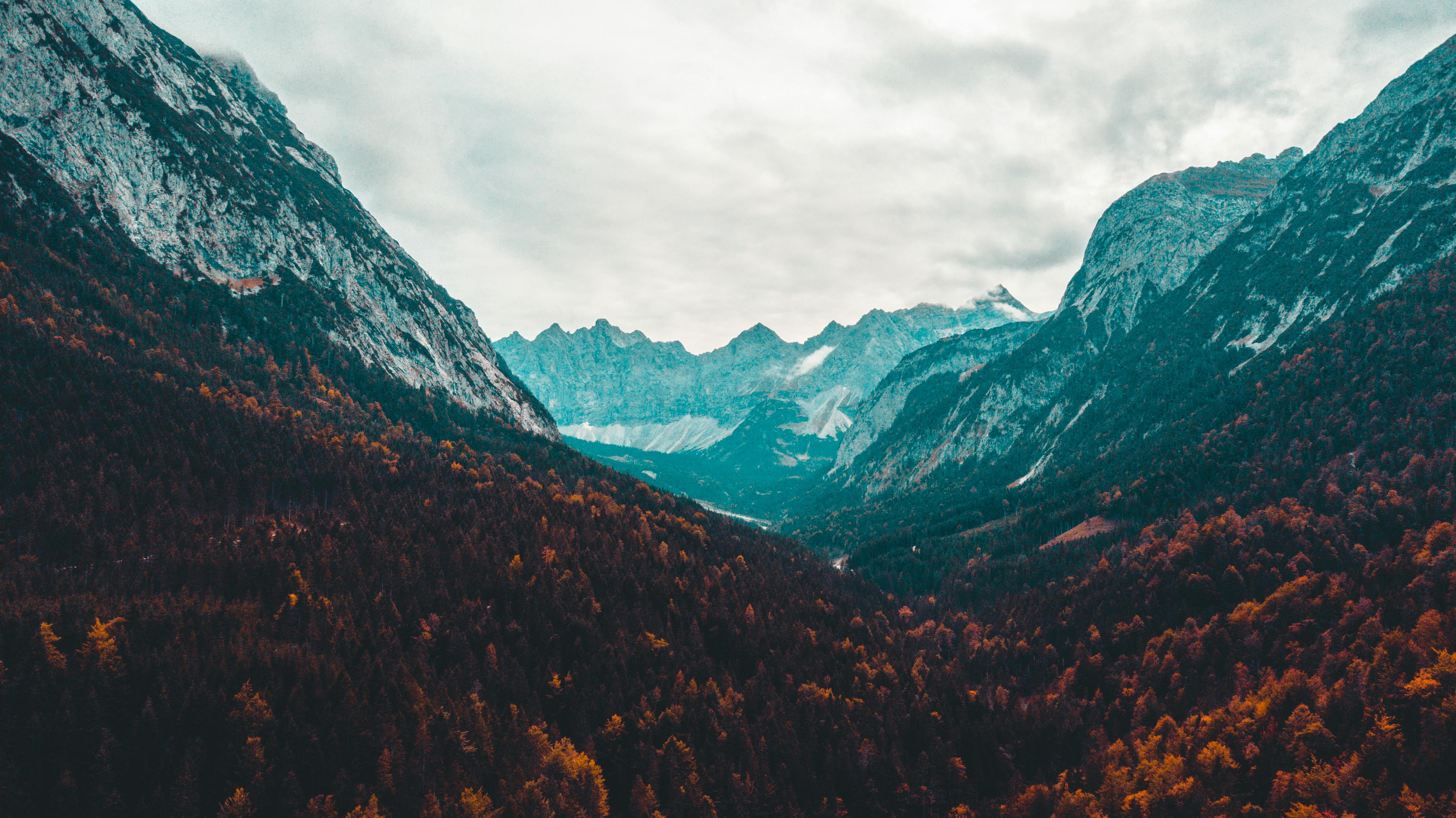 An overhead view of a valley, with mountains rising in the distance, the slopes covered in thick forests, and the sky in the distance is full of clouds. - free wallpaper image