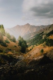 A small stream flows through a valley, with lush forests and grasslands on either side, towering mountains in the distance, and a cloudy sky. - free wallpaper image