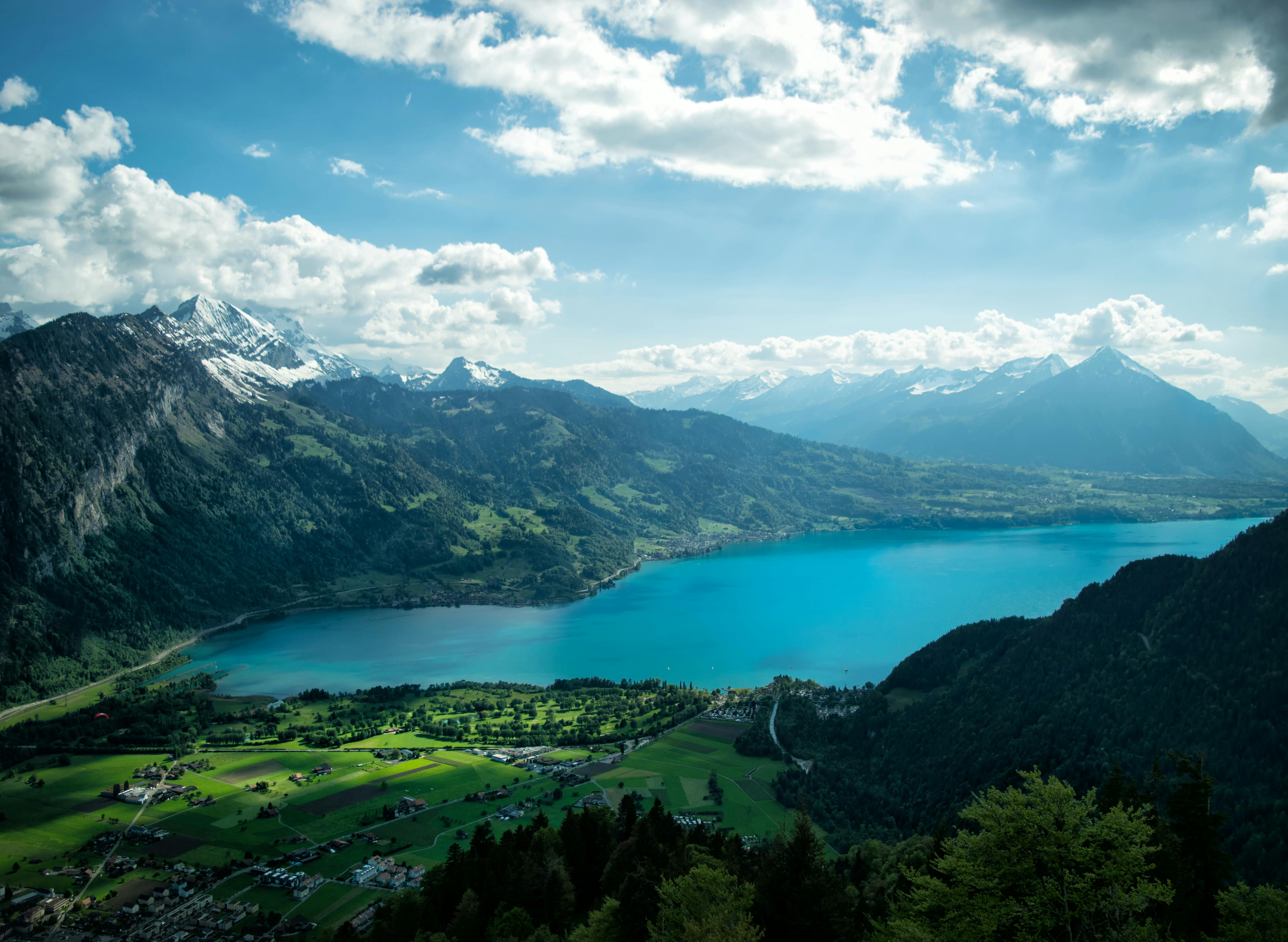 An aerial view of a valley with a winding turquoise lake surrounded by lush greenery and rolling mountains, snow-capped peaks glisten in the sunlight in the distance. - free wallpaper image