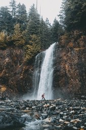 A person wearing a hat stands in front of a waterfall that cascades down from high rocks, surrounded by dense forest. - free wallpaper image