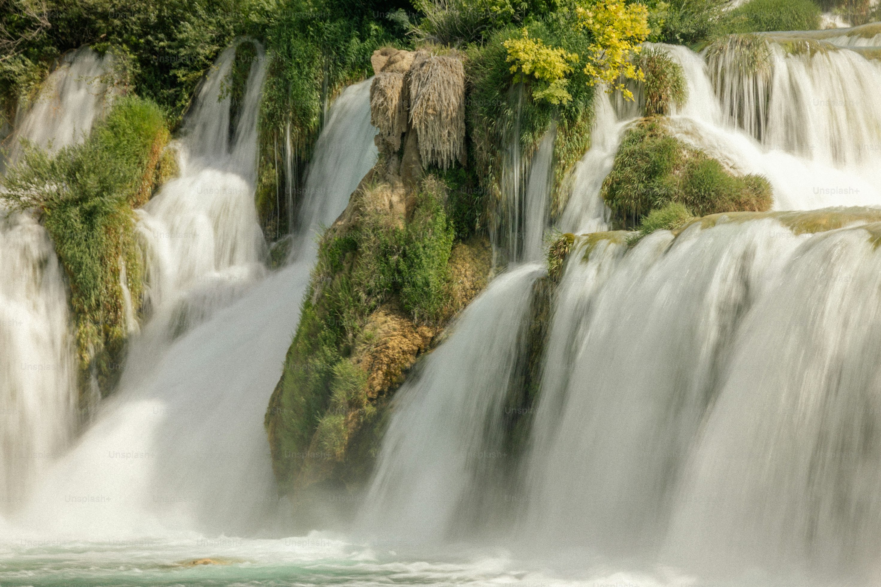 A waterfall cascades down rocky cliffs, the water flows rapidly, the spray is flying, surrounded by lush green vegetation. - free wallpaper image