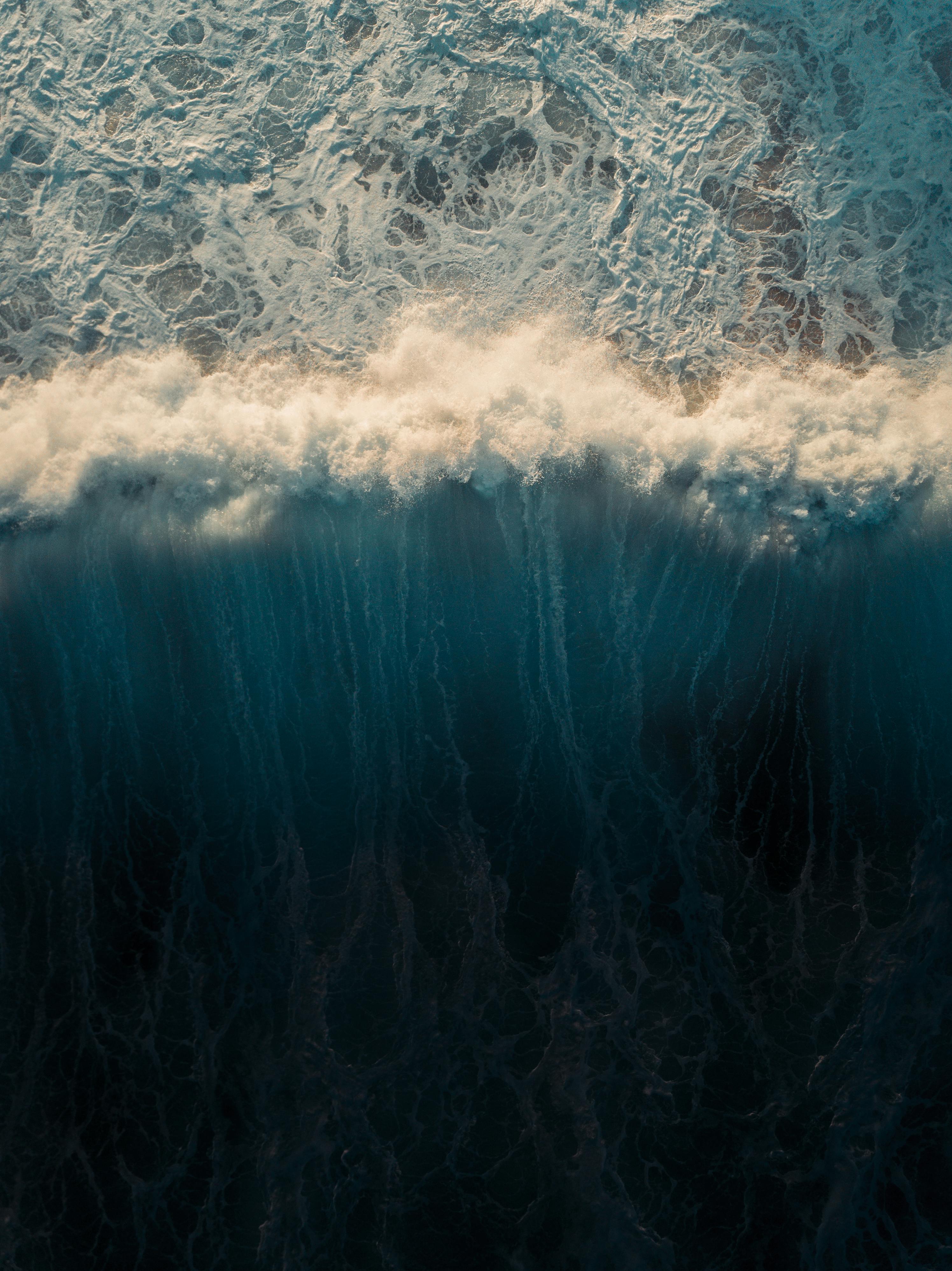 An aerial view of a wave crashing against the shore, forming white foam, the ocean water is deep blue. - free wallpaper image