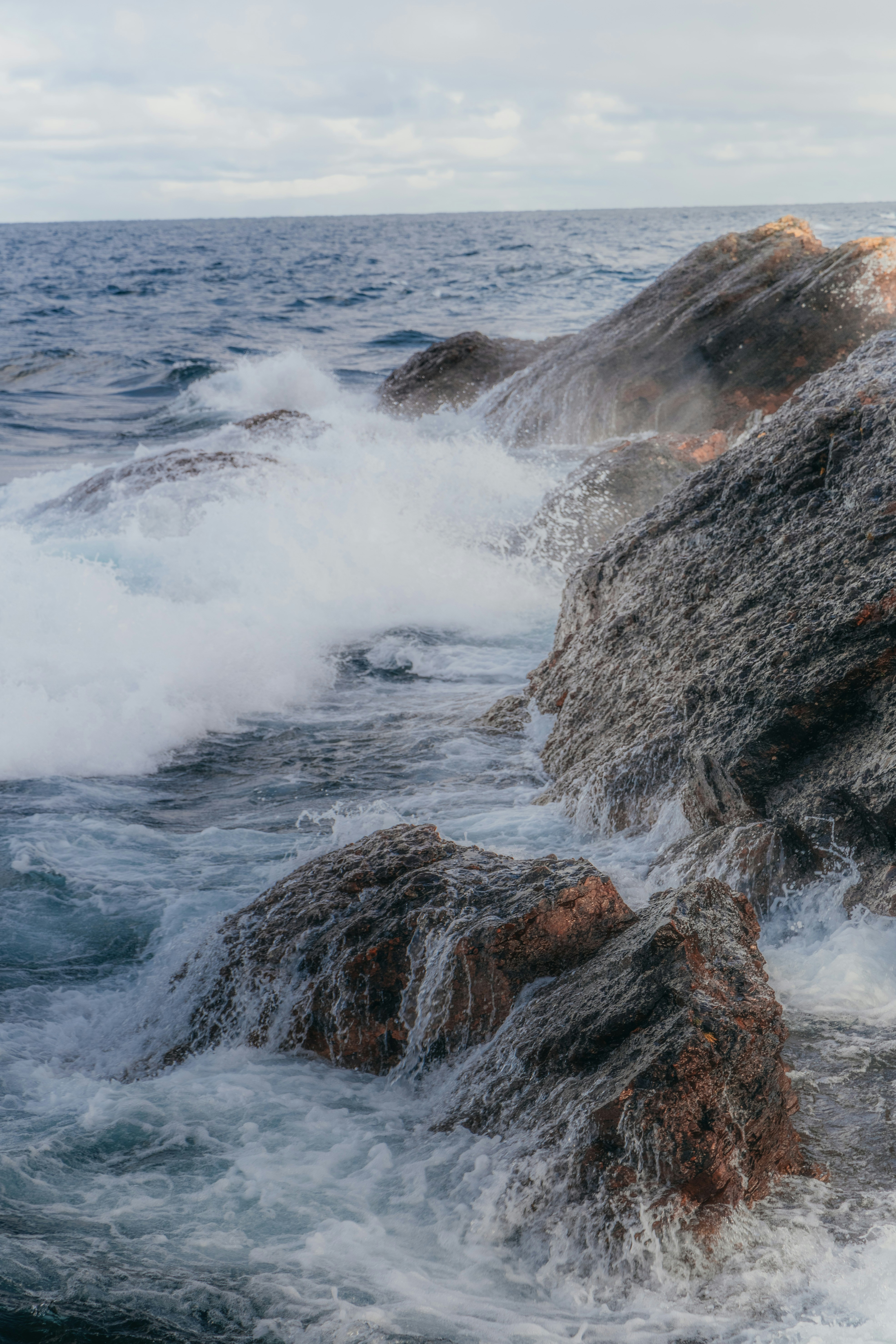 Waves crash against rocks, creating white foam, the sea is deep blue, and the sky is gray. - free wallpaper image