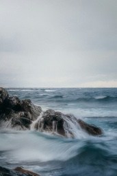 Waves crash against rocks, forming white spray, the sea surface is sparkling, and the sky is overcast. - free wallpaper image