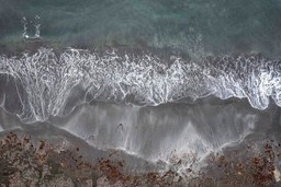 An aerial view of the coast with waves crashing against the shore, forming white foam. The ocean is a deep blue, and the coastline is clearly visible. - free wallpaper image