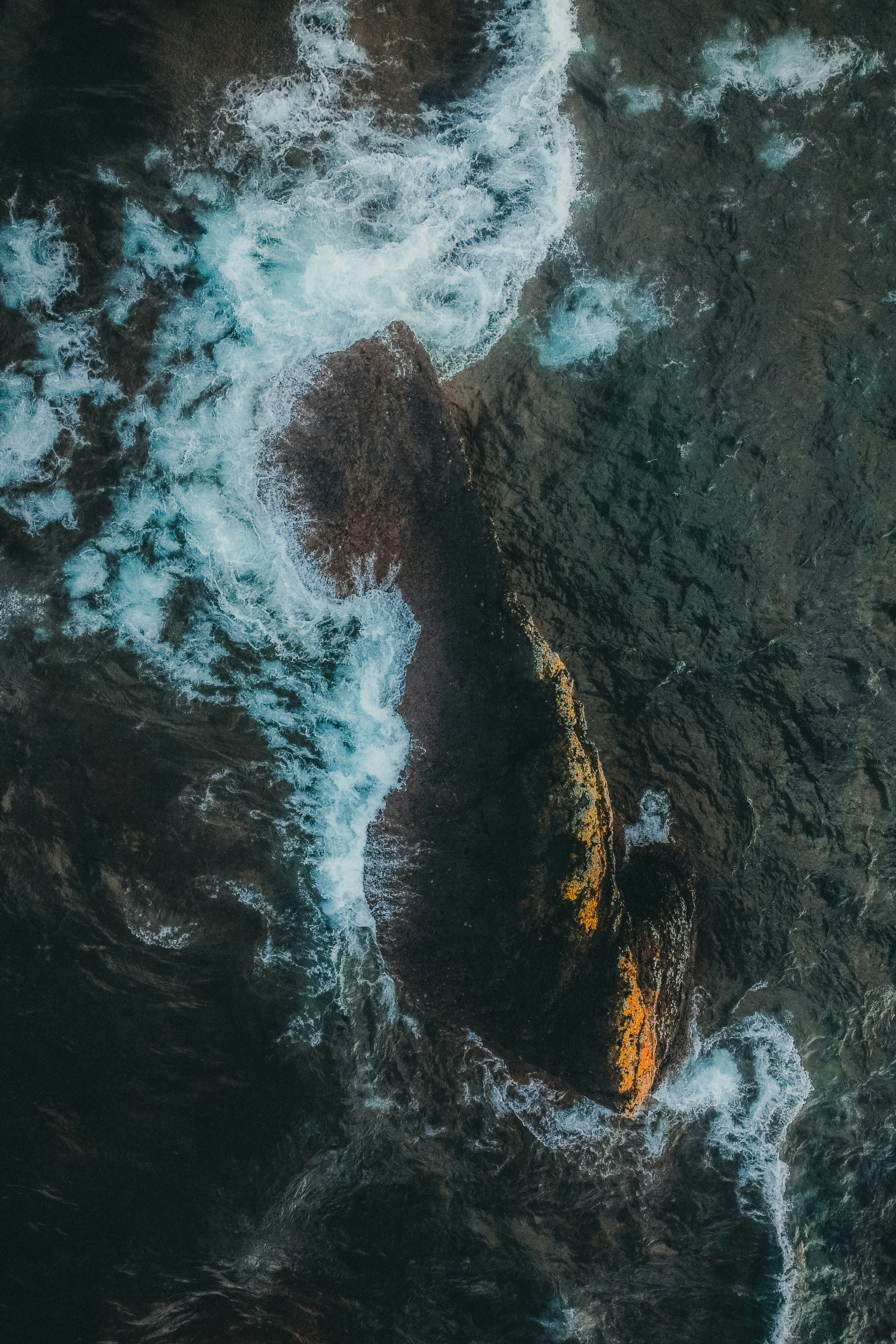 An aerial view of waves crashing against rocks on the shore, creating white foam, a dynamic scene. - free wallpaper image