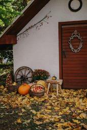 A white cabin with pumpkins, apples and a wooden wheel in front of it. There are many fallen leaves around, creating a strong autumn atmosphere. - wallpaper image