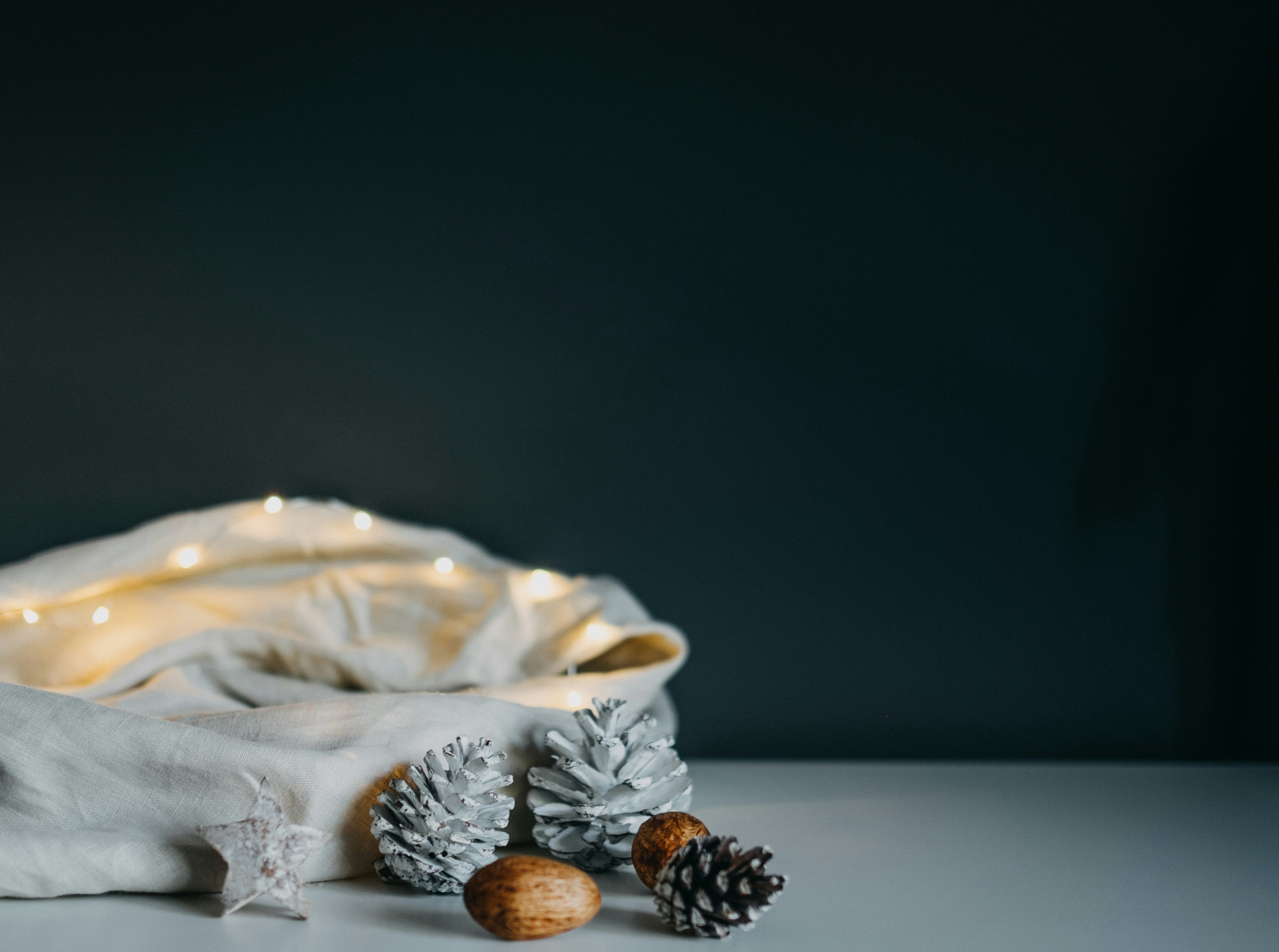 Two white pine cones are placed on a white cloth, next to some brown objects, there are also some small light bulbs on the cloth, and the background is a gray wall. - wallpaper image