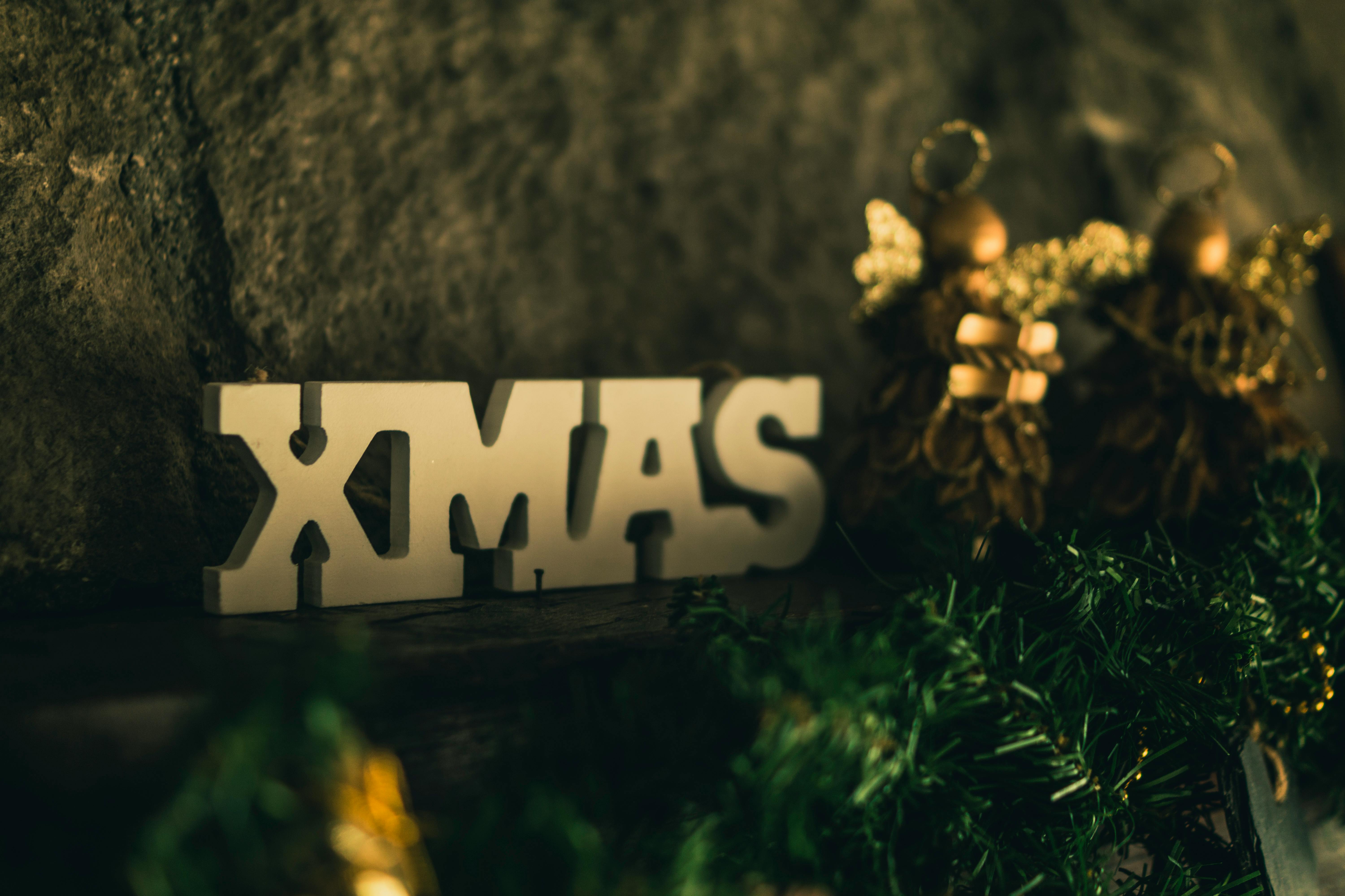 White "XMAS" letters are placed on a wooden table, with green Christmas decorations and gold decorations next to them, against a backdrop of rough stone walls. - wallpaper image