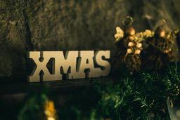 White "XMAS" letters are placed on a wooden table, with green Christmas decorations and gold decorations next to them, against a backdrop of rough stone walls. - wallpaper image