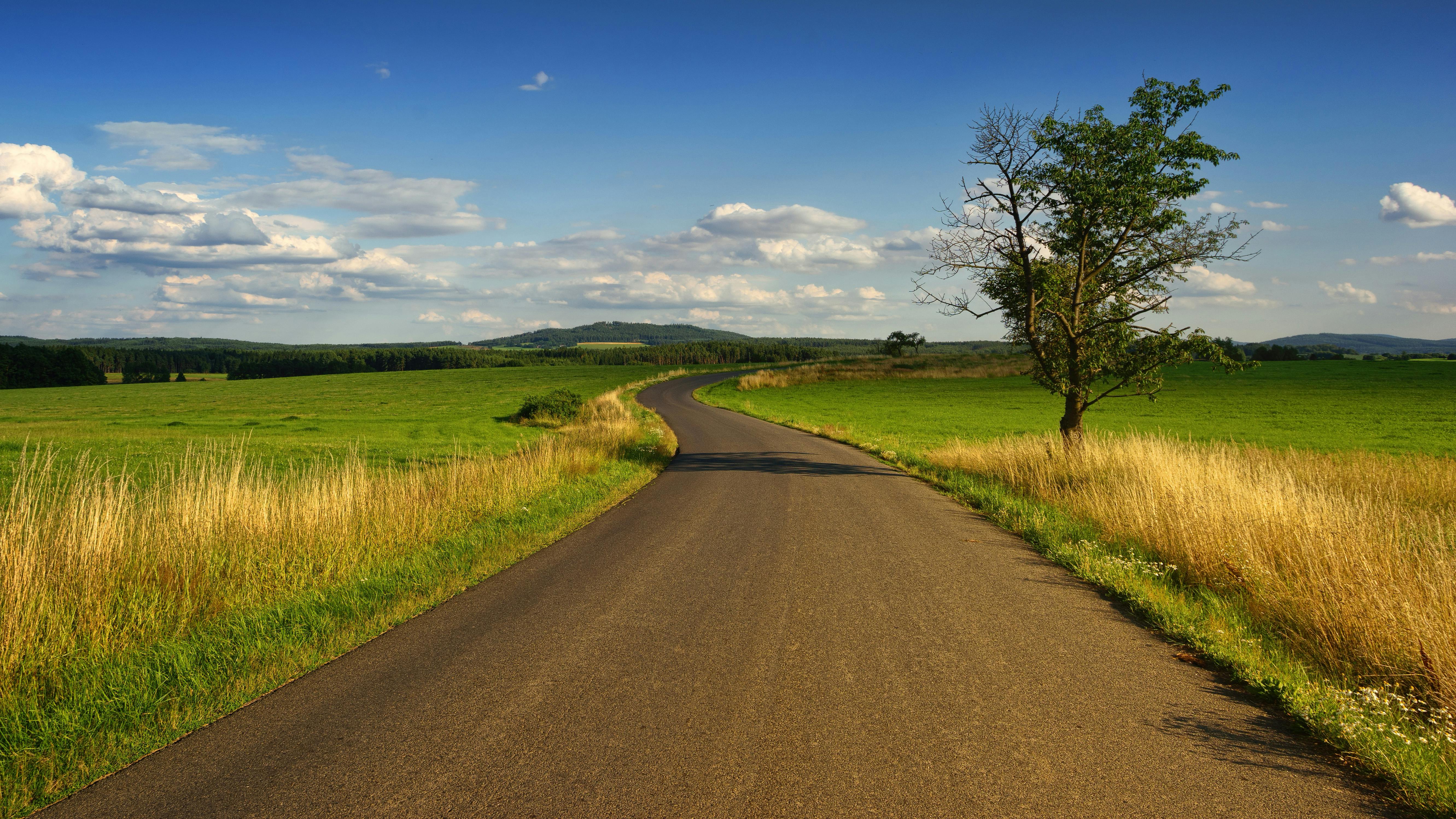 A winding asphalt road runs through a green field, with a blue sky and white clouds in the distance. - free wallpaper image