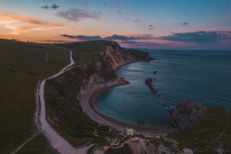 A winding road snakes through the hills towards a small cove on the coast, bathed in the golden light of sunset. - free wallpaper image