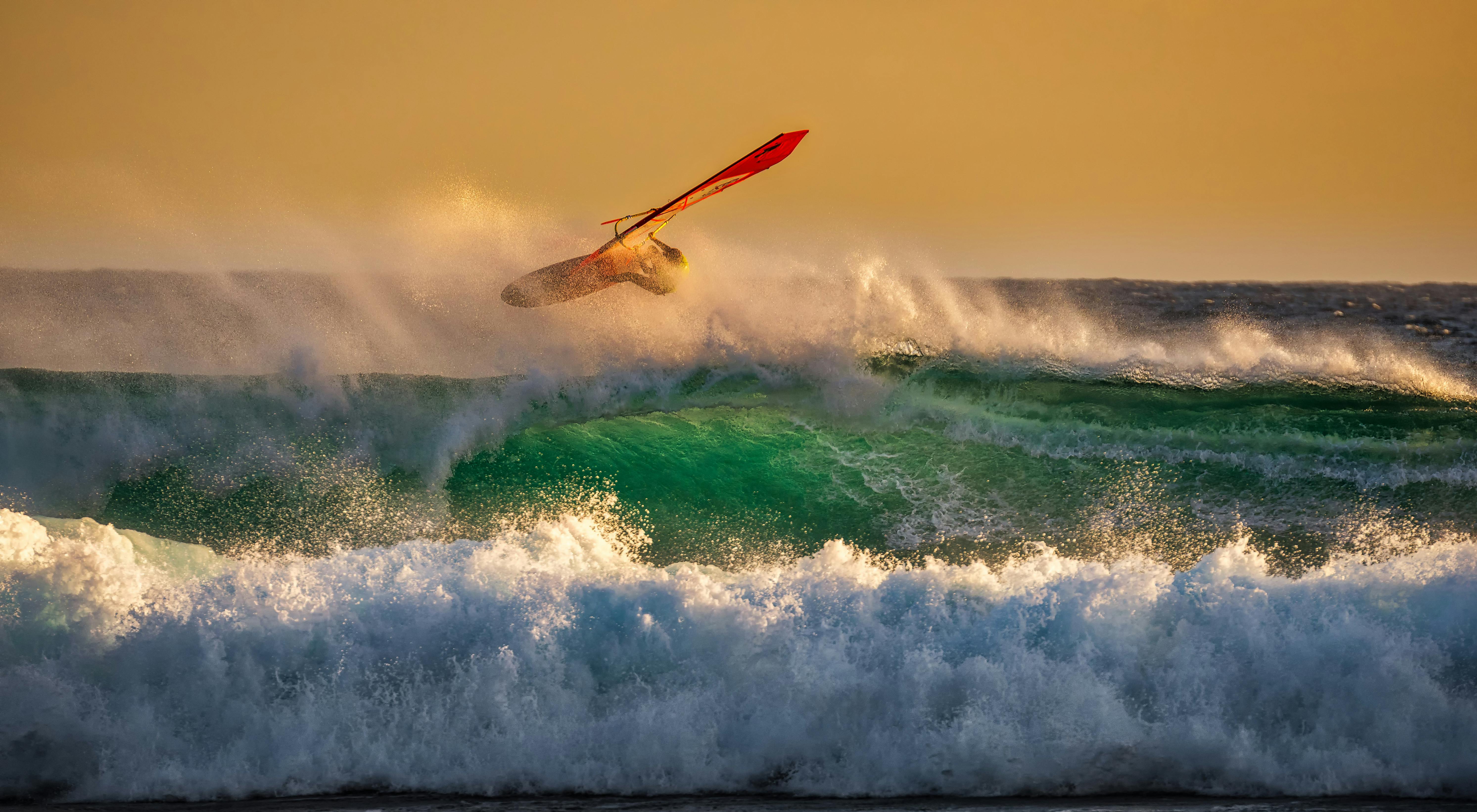 A windsurfer jumps over a wave, with a golden sunset and crashing waves in the background. - free wallpaper image