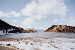 A lake in winter, the water is clear, the shore is covered with white snow, the distant mountains are covered with snow, the sky is clear and the clouds are white. - free wallpaper image