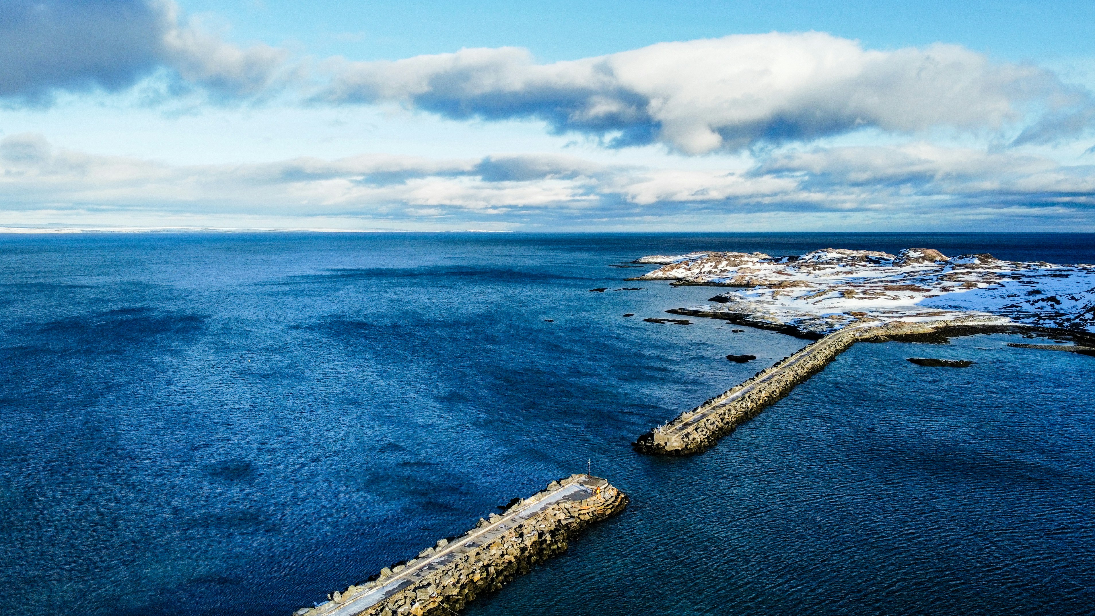 An aerial view of a long stone breakwater extending into the sea, the surface of the water is sparkling, the distant coastline is covered with snow, and the sky is filled with drifting clouds. - free wallpaper image