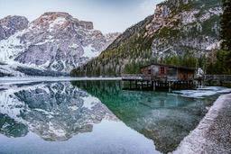 A wooden cabin stands on the calm lake, the water is clear and reflects the snowy mountains and surrounding trees. - free wallpaper image