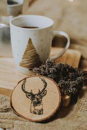 A wooden coaster with a deer design, next to a cup of hot drink and a bunch of dried flowers. - wallpaper image