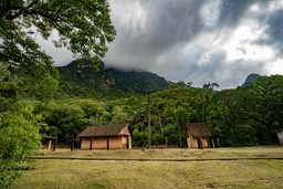 Several wooden houses are located at the foot of the mountain, surrounded by dense forests, with clouds in the sky. - free wallpaper image